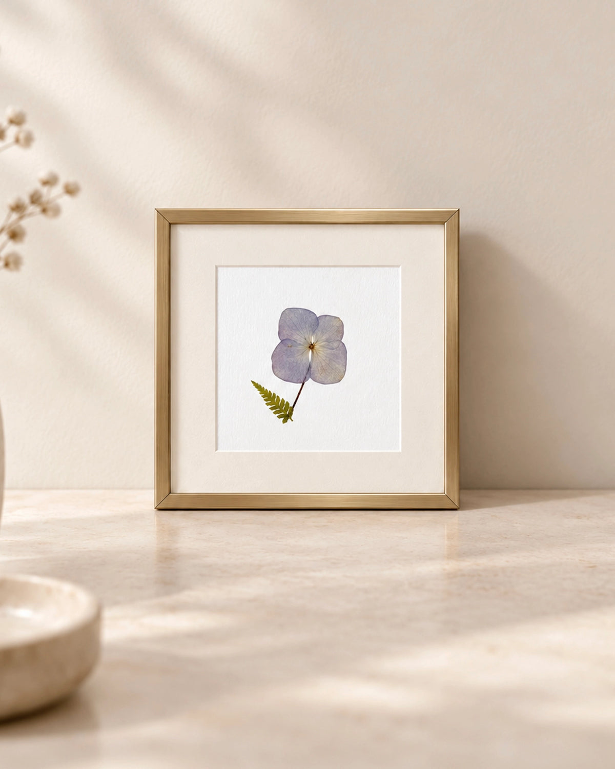 A framed botanical art print featuring a single light pressed Hydrangea botanical flower with a green fern leaf on a white mat, set in a gold frame on a light beige marble surface against a neutral beige wall.