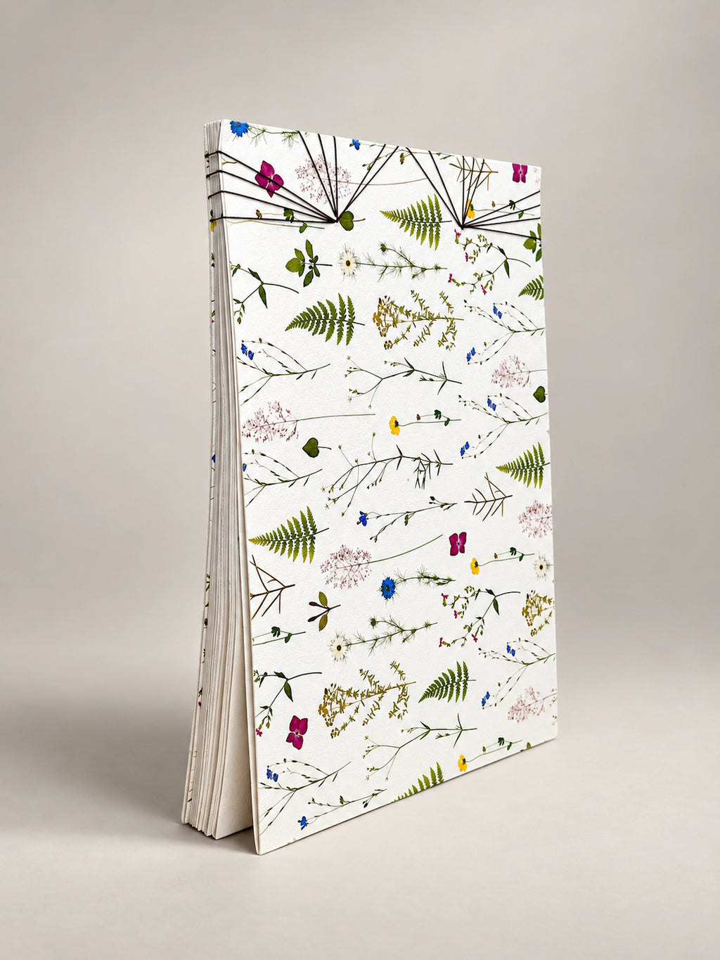 A tall, slim notebook or journal standing upright on a light gray background. The cover is white with an all-over botanical pattern of green ferns and small multicolored wildflowers, and the pages are visible along the left side. hand made in south west france