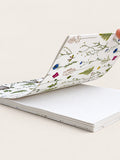 A hardcover notebook with a white cover decorated with delicate botanical illustrations, shown open with a hand lifting one page to reveal blank white interior pages. hand made in south west france