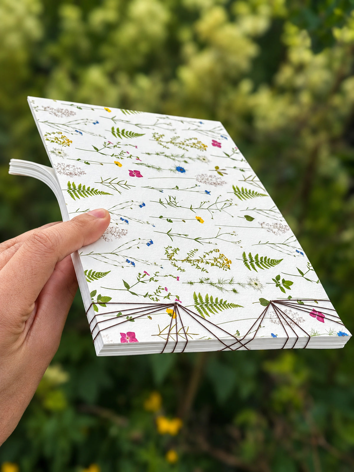 A hand holding a white hardcover journal decorated with a delicate green and multicolor pressed floral and botanical pattern, set against a blurred green outdoor background.