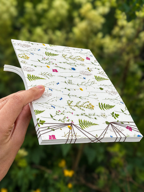 A hand holding a white hardcover journal decorated with a delicate green and multicolor pressed floral and botanical pattern, set against a blurred green outdoor background.