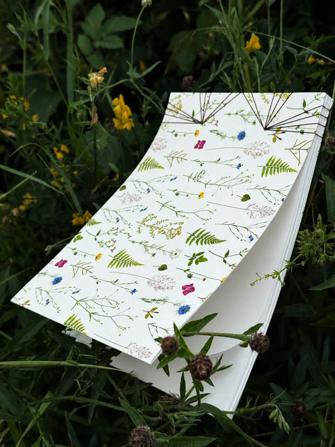 a premium handmade french journal featuring a delicate botanical pattern of pressed flowers and leaves, styled outdoors on green foliage and wildflowers.