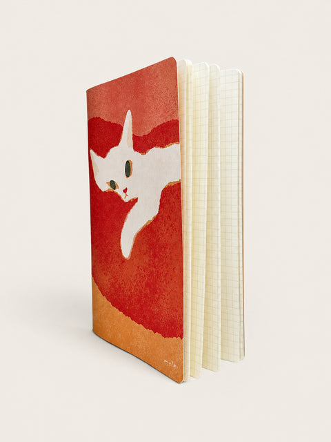 A notebook with a textured red and orange cover featuring a white cat illustration, standing upright to show its cream-colored grid paper pages.