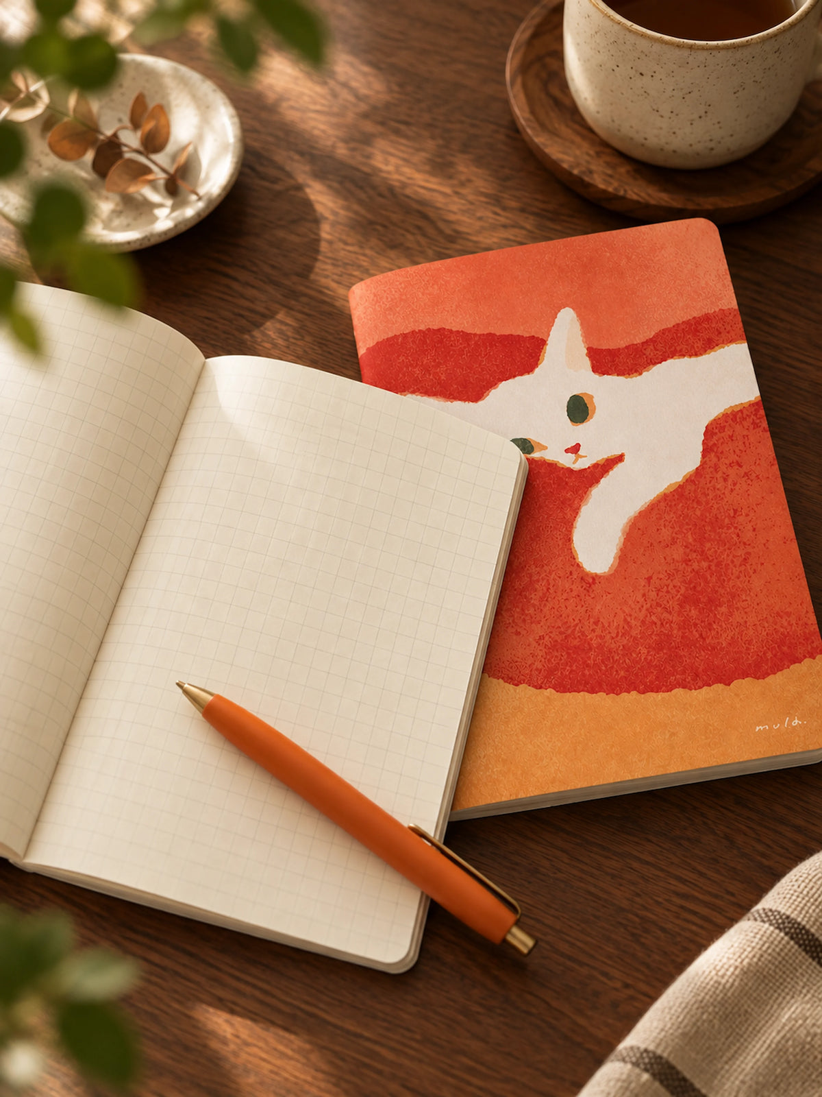 A styled flat-lay of a notebook with a colorful cat illustration on the cover, placed on a wooden table next to an open grid notebook, an orange pen, and a ceramic mug.