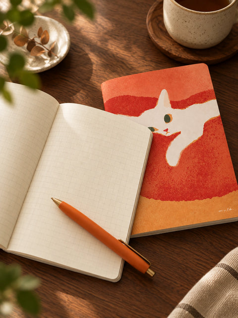 A styled flat-lay of a notebook with a colorful cat illustration on the cover, placed on a wooden table next to an open grid notebook, an orange pen, and a ceramic mug.
