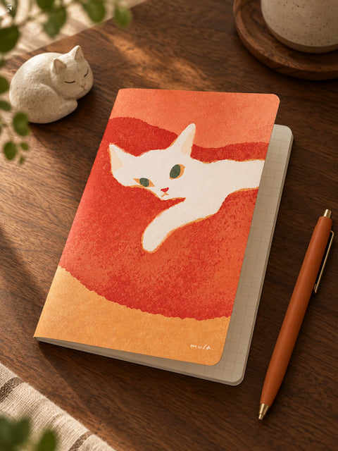 A notebook with a watercolor-style illustration of a white cat on an orange and red background, styled on a wooden desk next to a pen, a small cat figurine, and a ceramic cup.