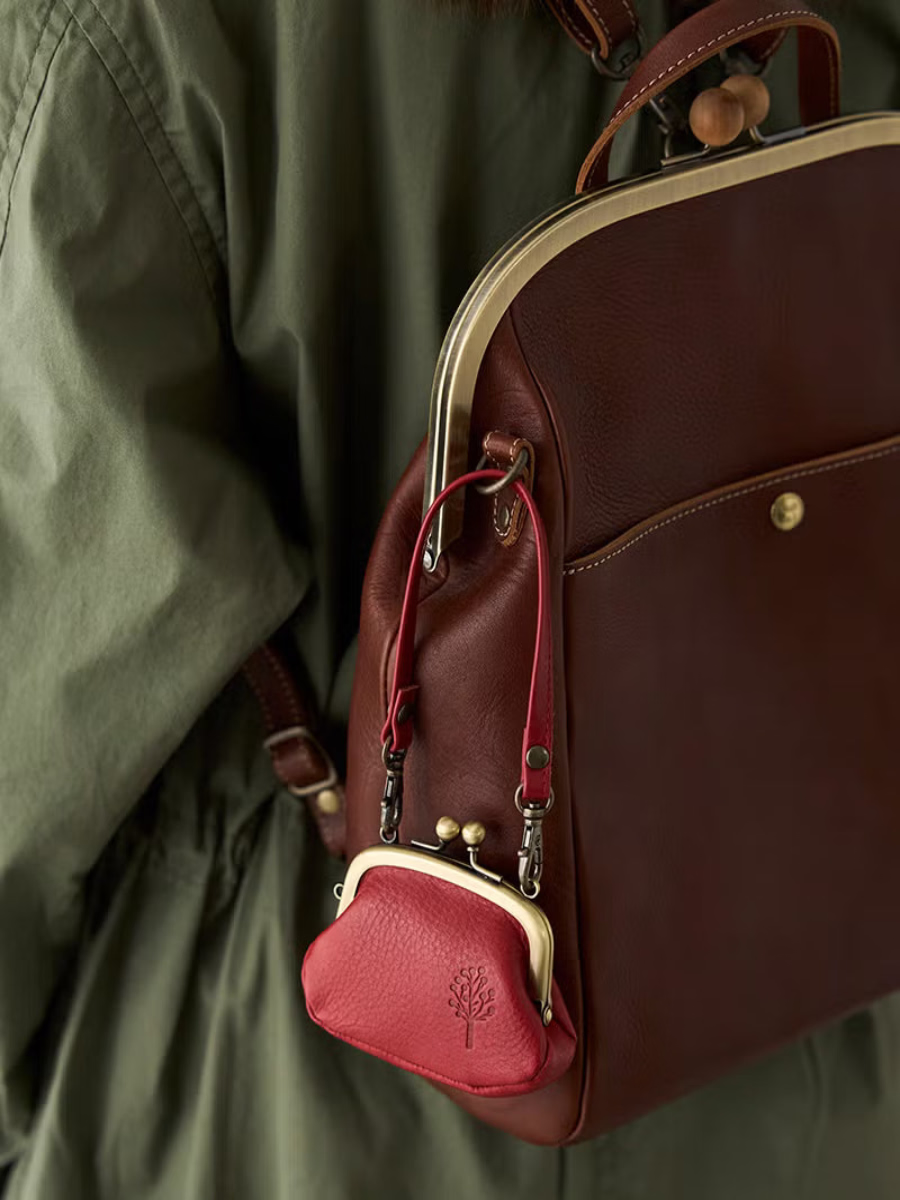 Deep red leather kiss lock coin purse clipped to the strap of a dark leather backpack