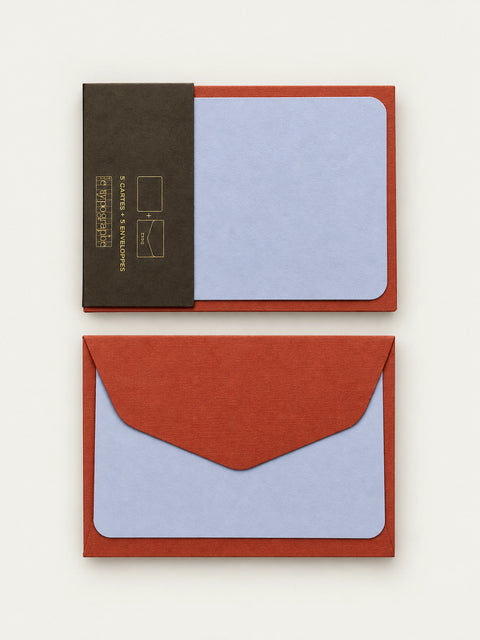 Lavender letterpress note card held in its charcoal paper band against a cordyline envelope, shown above a second envelope with a card behind the flap.