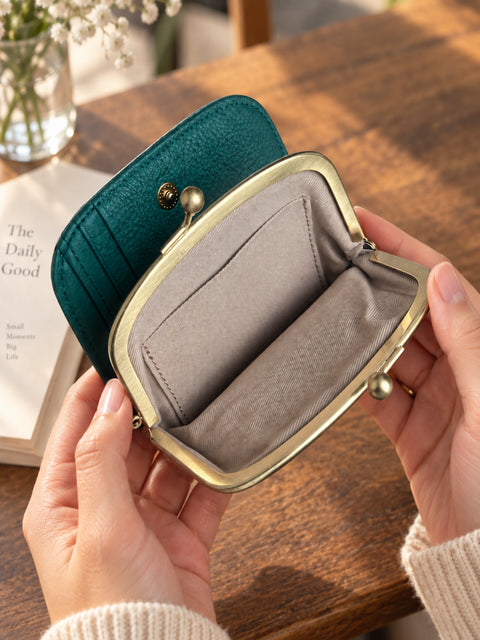 A teal green kiss-lock wallet with a gold-tone metal frame and a gray fabric interior, held open by two hands over a wooden table background.
