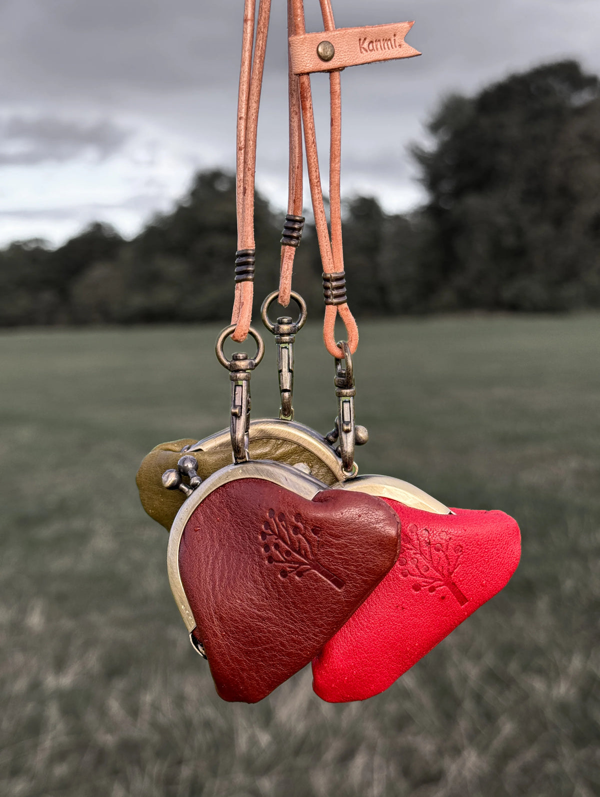 Three heart-shaped leather lockets hanging from tan leather straps outdoors in a grassy field. The lockets are in red, dark brown, and olive green, with brass clasps and a small leather tag that reads 'Kamino'.