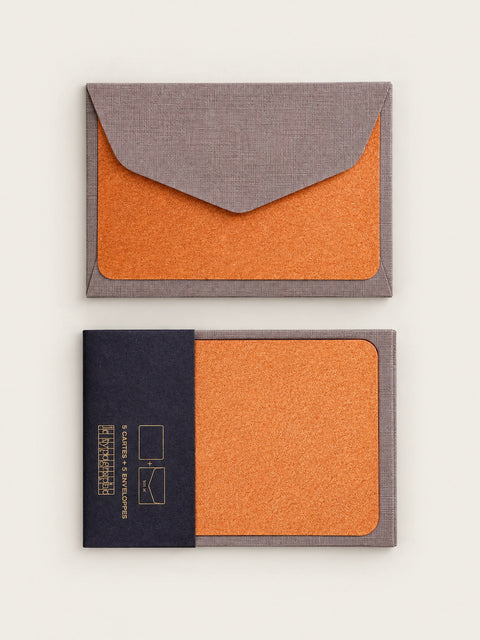 Copper letterpress note card tucked into a barley grey envelope, shown above a second card held in its charcoal paper band printed in gold.