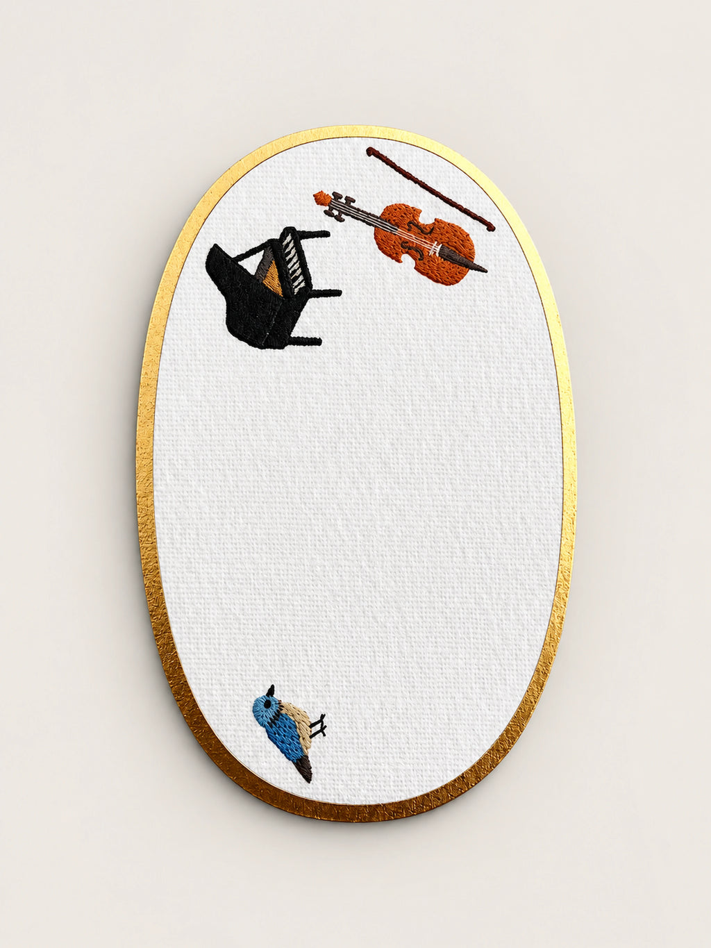 An oval embroidered card with a gold metallic border, featuring a white textured center embroidered with a black grand piano, an orange violin, a brown bow, and a small blue bird.