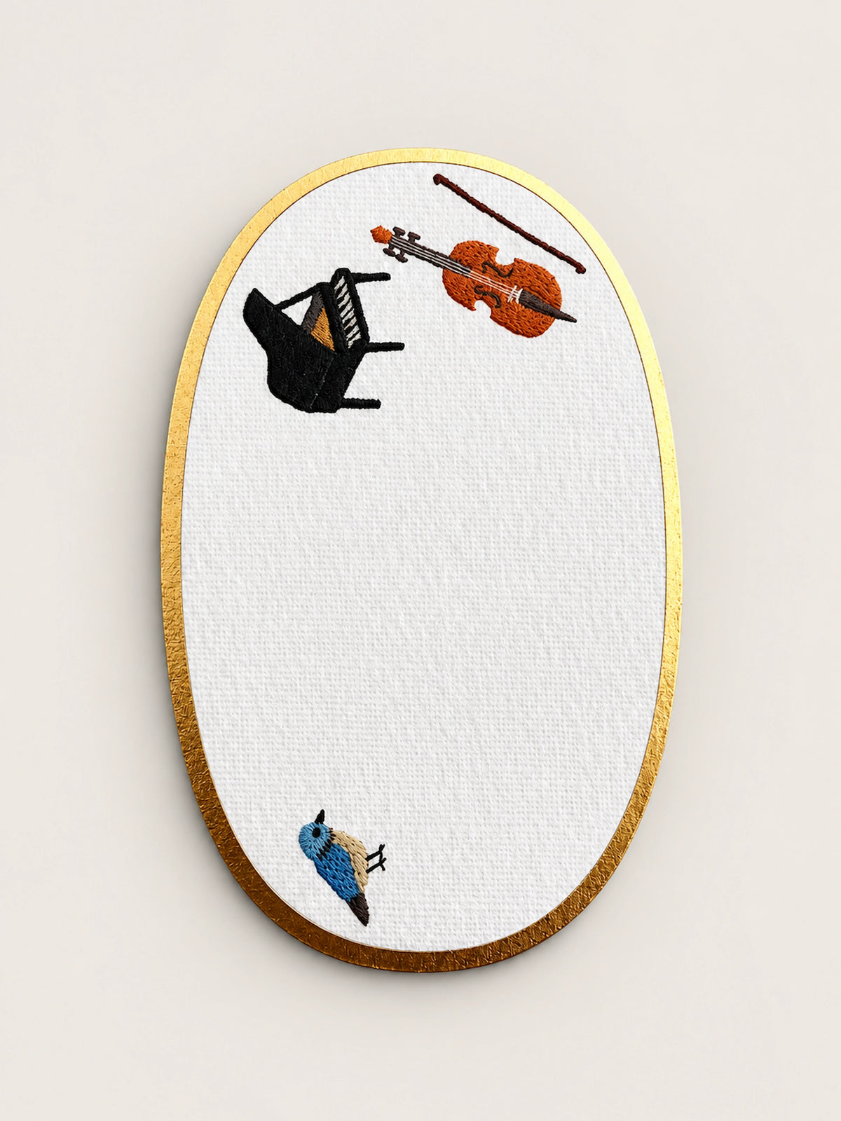 An oval embroidered card with a gold metallic border, featuring a white textured center embroidered with a black grand piano, an orange violin, a brown bow, and a small blue bird.