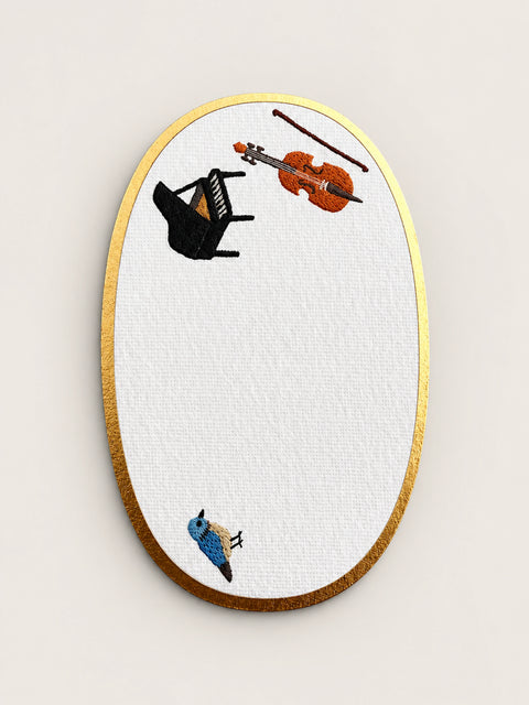 An oval embroidered card with a gold metallic border, featuring a white textured center embroidered with a black grand piano, an orange violin, a brown bow, and a small blue bird.