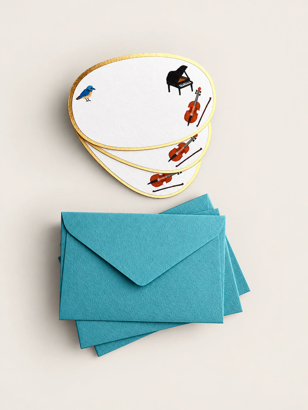 A set of white guitar-pick-shaped cards with gold borders and small illustrations of a bird, piano, cello, and violins, arranged above a stack of teal envelopes on a light beige background.