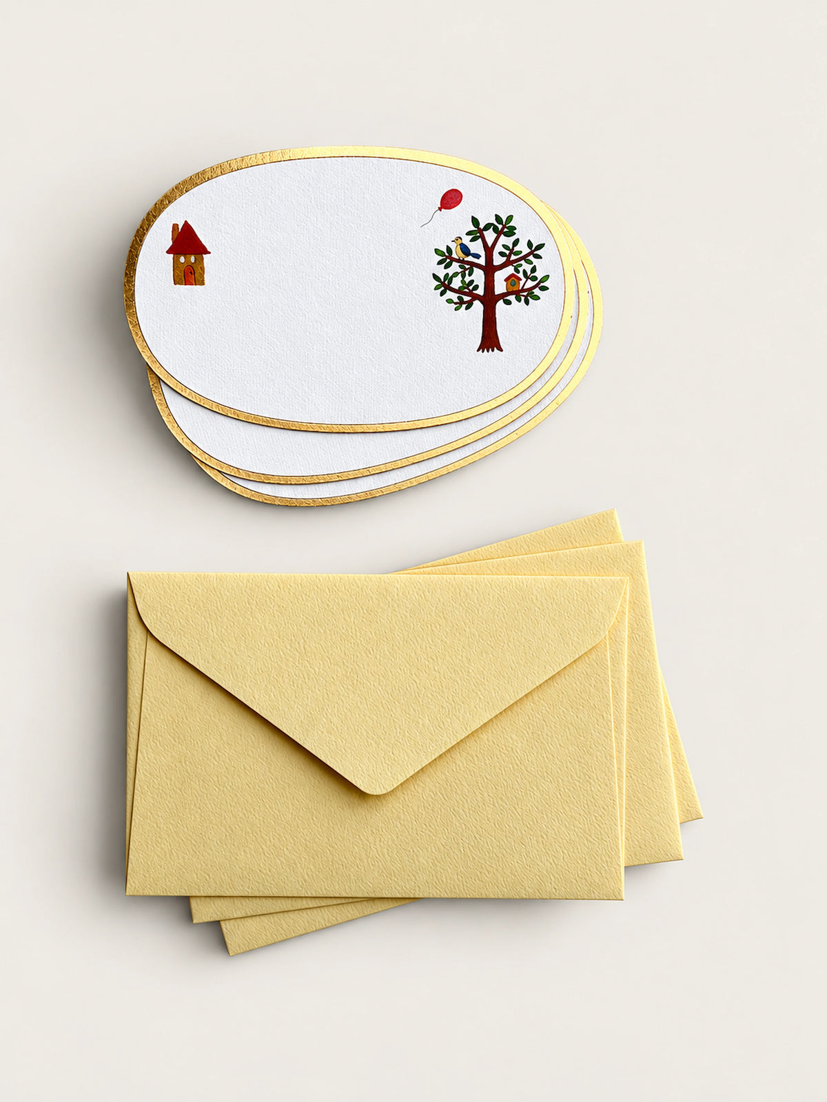 A set of oval white stationery cards with gold edges, printed with a small house and a tree with a red balloon, shown with matching pale yellow envelopes on a light beige background.