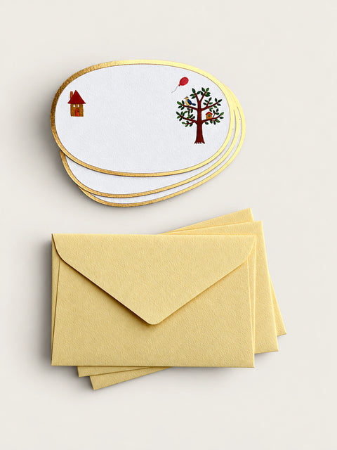 A set of oval white stationery cards with gold edges, printed with a small house and a tree with a red balloon, shown with matching pale yellow envelopes on a light beige background.
