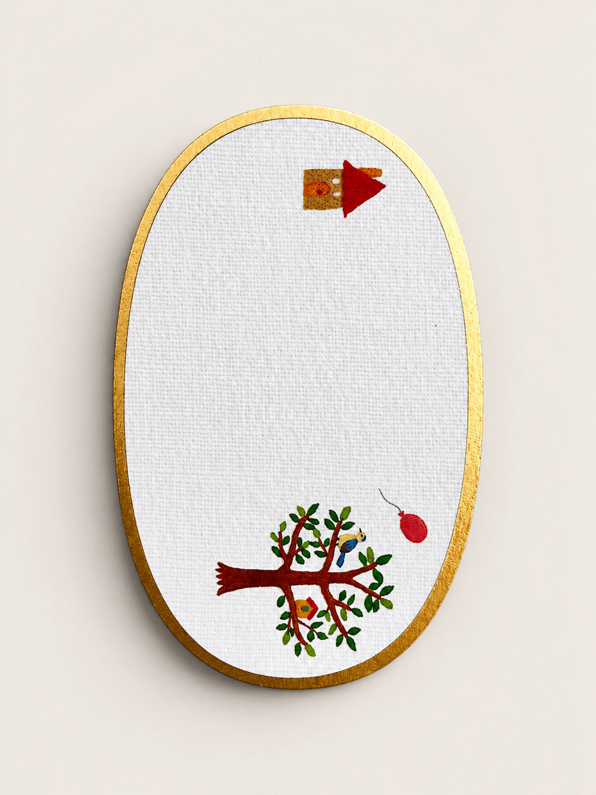 An oval embroidery card on white textured fabric with a gold embroidered border, featuring a stylized tree with green leaves and red branches, a small birdhouse, and a red hanging ornament.
