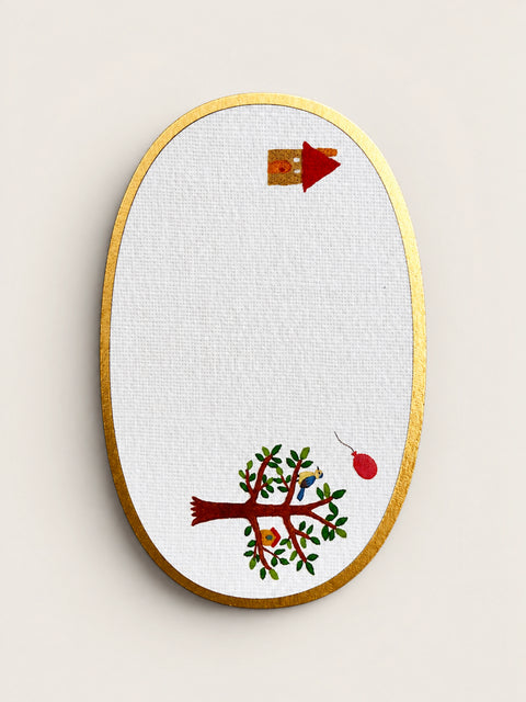An oval embroidery card on white textured fabric with a gold embroidered border, featuring a stylized tree with green leaves and red branches, a small birdhouse, and a red hanging ornament.