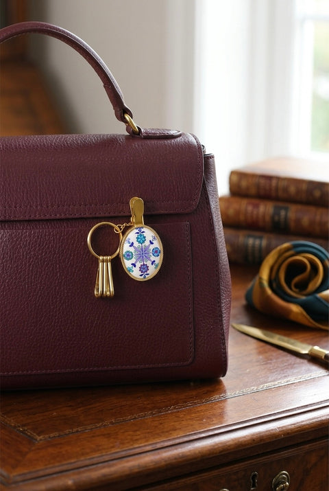 A burgundy leather handbag with a gold-tone key ring and decorative oval charm, resting on a polished wooden table with books and a scarf in the background.