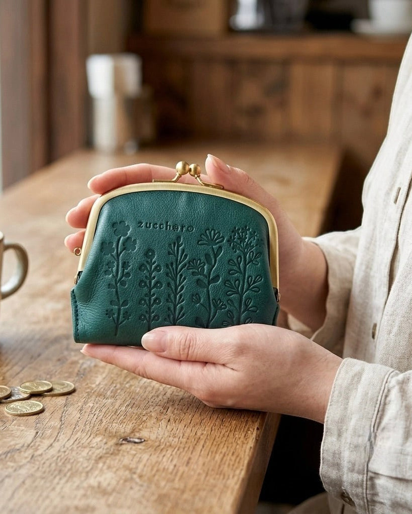 A person holds a small dark green kiss-lock wallet with embossed floral patterns and the brand name 'zucchero' on the front, set against a warm, rustic background with a wooden table, coffee mug, and coins.